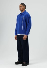 Blauw oversized poloshirt met lange mouwen, met witte tekst aan de zijkant. Donkere denim wijde pijpen broek en zwarte platform schoenen.