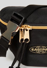 Sac Eastpak noir avec fermeture éclair dorée, doubles tirettes avec cordons noirs et une bandoulière noire réglable avec boucle métallique dorée.