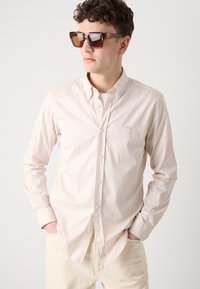Camicia button-up a righe beige con colletto, caratterizzata da righe bianche e marrone chiaro. Indossata con occhiali da sole marroni. I pantaloni non sono visibili.