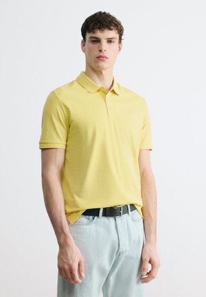 Junger Mann steht, trägt ein gelbes Polohemd mit kurzen Ärmeln, das in eine hellblaue Jeans mit schwarzem Gürtel gesteckt ist.