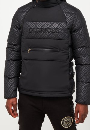 Veste matelassée noire avec motif géométrique sur les manches et la poitrine, "GLORIOUS" brodé sur le devant, poche zippée, portée avec un pantalon noir affichant un emblème circulaire.