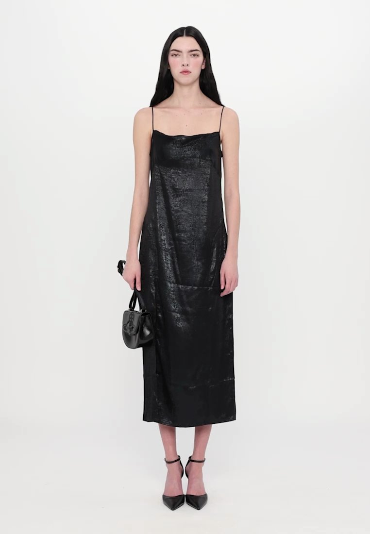 Robe nuisette en satin noir avec de fines bretelles, coupe droite et fente sur le côté. Le mannequin tient un petit sac à main noir et porte des talons aiguilles.