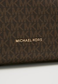 Tissu marron texturé avec un motif répétitif marron foncé "MK" et des lettres métalliques dorées formant "MICHAEL KORS" sur une surface plane.