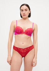 Roze push-up beha met kantdetails, voorzien van een strikje in het midden en verstelbare bandjes. Rode kanten string met bloemmotieven op de tailleband.