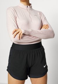 Světle růžový dlouhý rukáv s límcem na zip a šedým logem Nike, spojený s černými sportovními šortkami s elastickým pasem a detailním logem.
