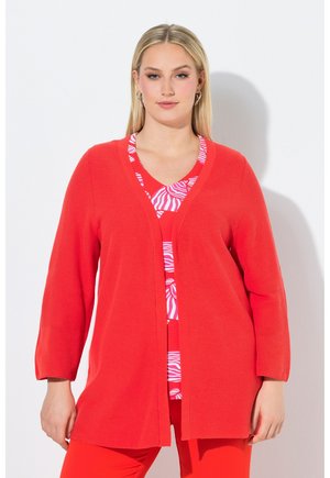 Donna che indossa un cardigan rosso acceso aperto davanti su una maglia rossa e bianca a motivo, abbinato a pantaloni rossi, in piedi davanti a uno sfondo bianco.