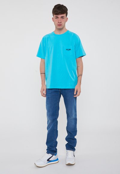 Maglietta turchese con colletto rotondo e taschino, abbinata a jeans blu e sneaker bianche con dettagli blu. Design semplice e casual.