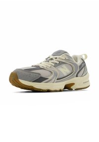 Zapatilla deportiva en tonos gris y beige, confeccionada con materiales de malla y ante, que presenta una suela de goma, cordones y el logo de New Balance en el lateral.