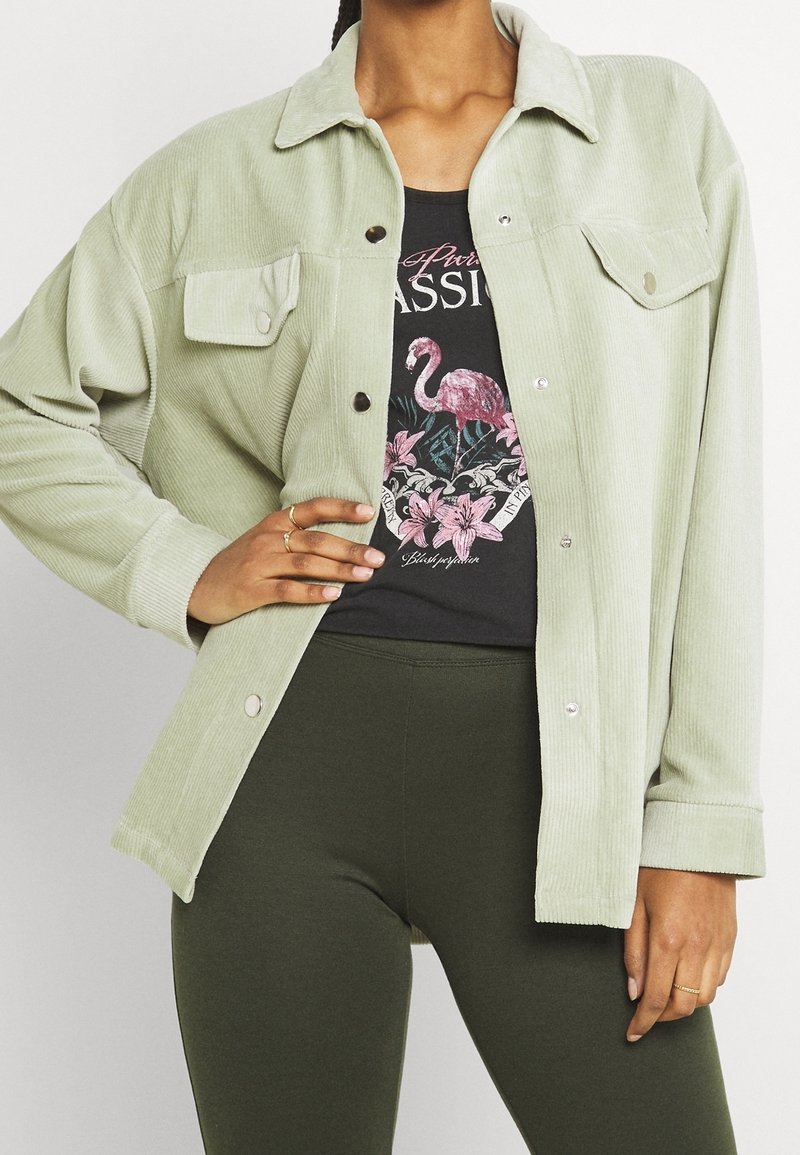 Giacca in corduroy verde chiaro con colletto, bottoni a pressione e due tasche sul petto. Indossata sopra una t-shirt nera con grafica e leggings verdi.