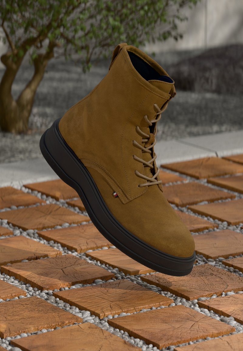 Bottine en daim marron avec semelle noire, bout rond et design à lacets, avec une petite étiquette logo sur le côté.