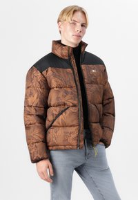 Chaqueta tipo puffer marrón con patrón floral, mangas negras, cuello alto, cierre frontal y bolsillos laterales. Textura suave con un aspecto acolchado.