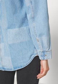 Světle modrá džínová košile s patchworkovým designem. Bytové manžety na knoflíky a zakřivený lem. Materiál je měkký s mírně texturovaným povrchem.