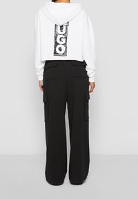 Bílá zkrácená mikina s kapucí s černým logem na zádech, doplněná volnými černými cargo kalhotami s bočními kapsami a měkkou texturou.