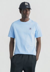 Ανοιχτό μπλε βαμβακερό t-shirt με κοντά μανίκια και κυκλικό λογότυπο Converse. Συνδυασμένο με μαύρο παντελόνι και μπλε καπελάκι με ρίγες.