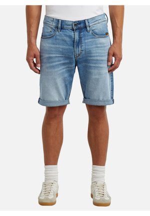 Man draagt lichtblauwe opgerolde denimshorts, witte sokken en witte vetersneakers, staand tegen een effen achtergrond.
