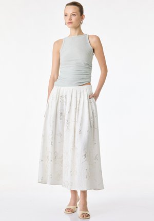 Kobieta ubrana w bezrękawnik w jasnoszarym kolorze, długą spódnicę w biały kwiatowy wzór oraz beżowe sandały na obcasie z odkrytymi palcami, stojąca z jedną ręką w kieszeni.