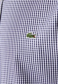 Camicia a motif gingham blu navy e bianco con un piccolo logo di coccodrillo verde ricamato sull'area del petto. Tessuto in cotone con una texture liscia.