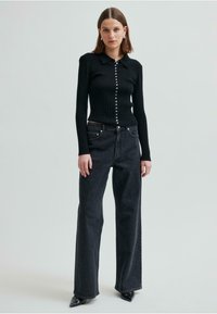 Svart ribbad långärmad tröja med krage och knäppning framtill, kombinerad med hög midja, vida mörka jeans och spetsiga svarta skor.