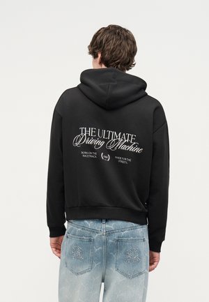 Άτομο που φοράει μαύρο φούτερ με κουκούλα με το κείμενο "The Ultimate Driving Machine" στην πλάτη, συνδυασμένο με ανοιχτού μπλε κεντημένο τζιν, γυρισμένο από πίσω.