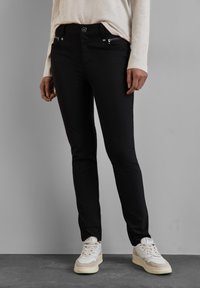 Osoba oblečená v černých úzkých kalhotách, béžovém dlouhém tričku a bílých teniskách, stojící na tmavé podlaze před šedým pozadím.