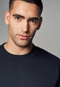 Mörkblå sweatshirt med ribbad rund halsringning. Tyget verkar mjukt med en slät yta. Modellen har kort, välansad frisyr.