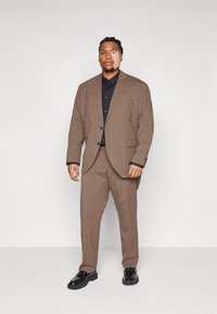 Brun rutad kostym med jacka med notch-lapel och matchande byxor, kombinerad med en svart skjorta med knappar och blanka svarta skor.
