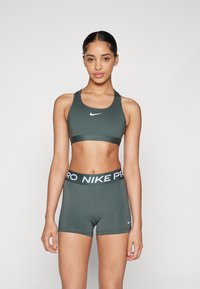 Žena u tamnozelenom Nike sportskom grudnjaku i odgovarajućim Nike Pro kratkim hlačama, stoji naslonjena na jednostavnu svijetlu pozadinu.