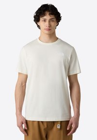 Cream cotton t-shirt with a crew neck, short sleeves, featuring a small white logo on the left chest, paired with brown pants.