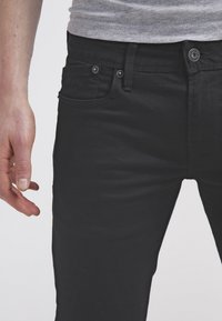 Černé džíny z denimu s úzkým střihem, designem pěti kapes, kovovým knoflíkem a nýty a jemnou strukturou. Ruka v těsné blízkosti.