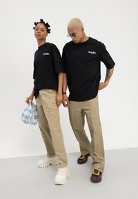 Zwei Personen tragen schwarze "Memorize"-T-Shirts und beige Hosen und stehen vor einem weißen Hintergrund, eine hält einen blau-weißen karierte Hut in der Hand.