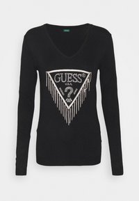 Musta, pitkähihainen neule V-aukkosella, jossa on kolmiomainen strassilogo, jossa hapsureunus alareunassa ja "GUESS®"-teksti.