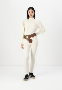 Pulover crem cu arcuri, cu mâneci bufante, asortat cu blugi skinny bej deschis și o curea lată maro cu accesorii circulare.