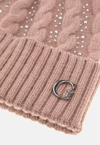 Gorro de punto en rosa claro con textura de trenzado, acentuado con tachuelas metálicas y un charm de logo plateado. Presenta un detalle de puño acanalado.