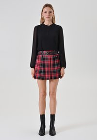 Schwarzes Langarmoberteil mit einem Schlüssellochausschnitt, kombiniert mit einem rot-schwarzen karierte Plisseerock mit einem Gürtel. Schwarze Knöchelboots runden das Outfit ab.