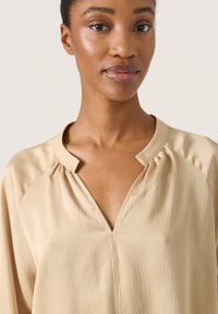 Beige bluse med V-udskæring, lavet af tekstureret stof, med raglanærmer og en samlet detalje ved halsudskæringen.