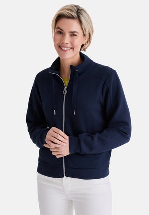 Smiling woman with short blonde hair wearing a navy zip-up jacket and white pants, standing against a plain background.