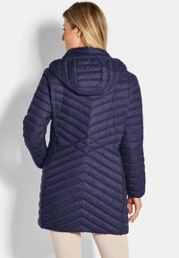 Vrouw met blond haar in een marineblauwe gewatteerde jas met capuchon, staand met haar rug naar de camera tegen een witte achtergrond.