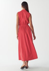 Czerwona, bezrękawnikowa sukienka maxi z uwydatnioną talią, luźnym krojem i pełnym długością. Ma kołnierz oraz gładką teksturę materiału.