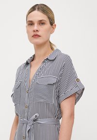 Vestido tipo camisa riscado em azul-marinho e branco. Apresenta mangas curtas, bolsos com botão, gola e uma cintura atada com uma textura de tecido suave.
