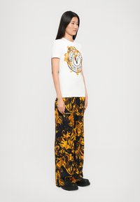 Sieviete stāv, ģērbusies baltā Versace T-kreklā ar zelta logo, melnās platās biksēs ar zeltainu baroka rakstu un melnās platformas kurpēs.