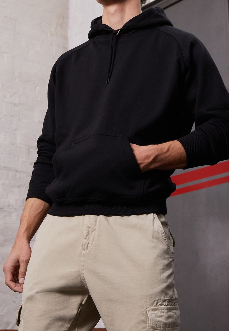 Homme portant un sweat à capuche noir avec poche avant et un pantalon cargo beige, main dans la poche, contre un mur clair à texture.