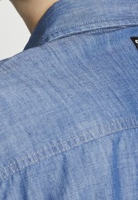 Bakåtrynan av en ljusblå jeansskjorta med synlig sömnad och en liten svart tygetikett.