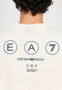 Cream t-shirt featuring a graphic print of three circular logos labeled "E," "A," and "7," with black text and a smooth texture.
