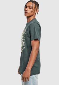 Mörkgrön t-shirt med kort ärm som har en grafisk tryck av en mumie, röda detaljer och text på framsidan, tillverkad av mjukt bomullsmaterial.