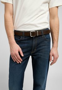 Brun läderbälte med en silverfärgad metallspänne, bärs över mörkblå jeans, med en enkel vit t-shirt synlig ovanför bältet.