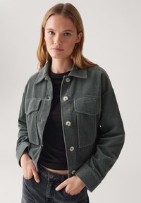 Donkergroene corduroy jas met een knoopsluiting aan de voorkant, twee borstzakken en een kraag; textiele stof met zichtbare ribbels.