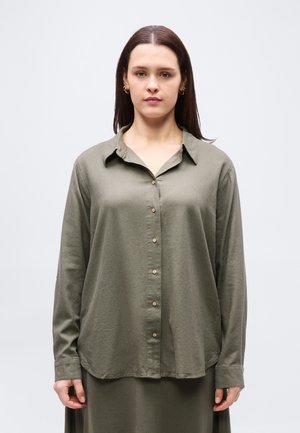 Žena s rovnými hnedými vlasmi má na sebe voľnú olivovo zelenú košeľu na gombíky a zodpovedajúcu sukňu, stojaca pred jednoduchým svetlosivým pozadím.