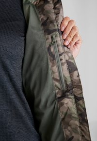 Main tenant une veste camouflage ouverte révélant une poche intérieure zippée et une doublure en maille verte sur une chemise sombre texturée.