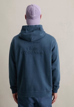 Marineblauwe hoodie met geborduurde tekst op de achterkant, met een relaxte pasvorm en een kangoeroezak; gecombineerd met een lichtpaarse pet.