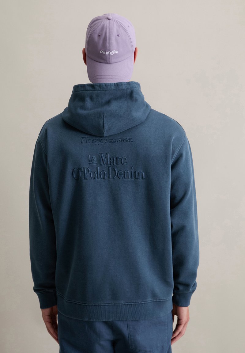 Marineblauwe hoodie met geborduurde tekst op de achterkant, met een relaxte pasvorm en een kangoeroezak; gecombineerd met een lichtpaarse pet.